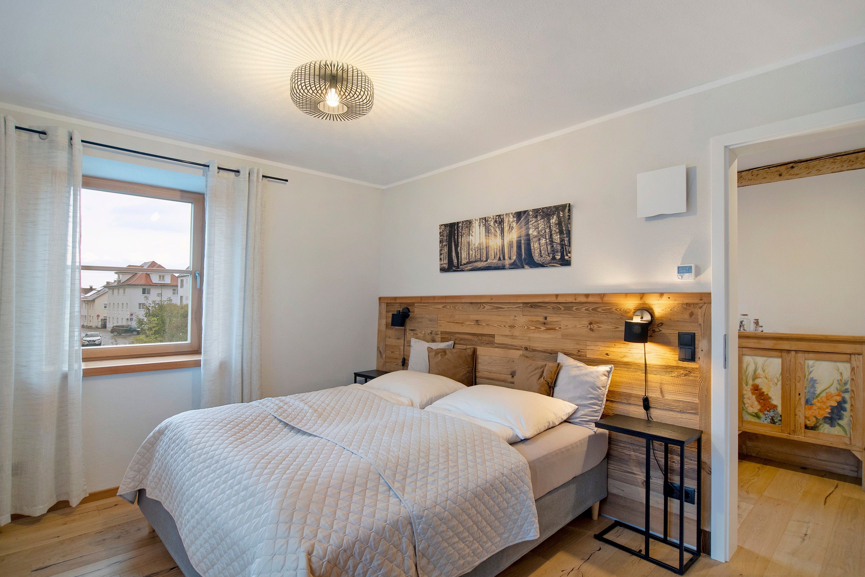 A bedroom with a bed, a painting, a nightstand, a lamp, and a window with curtains.