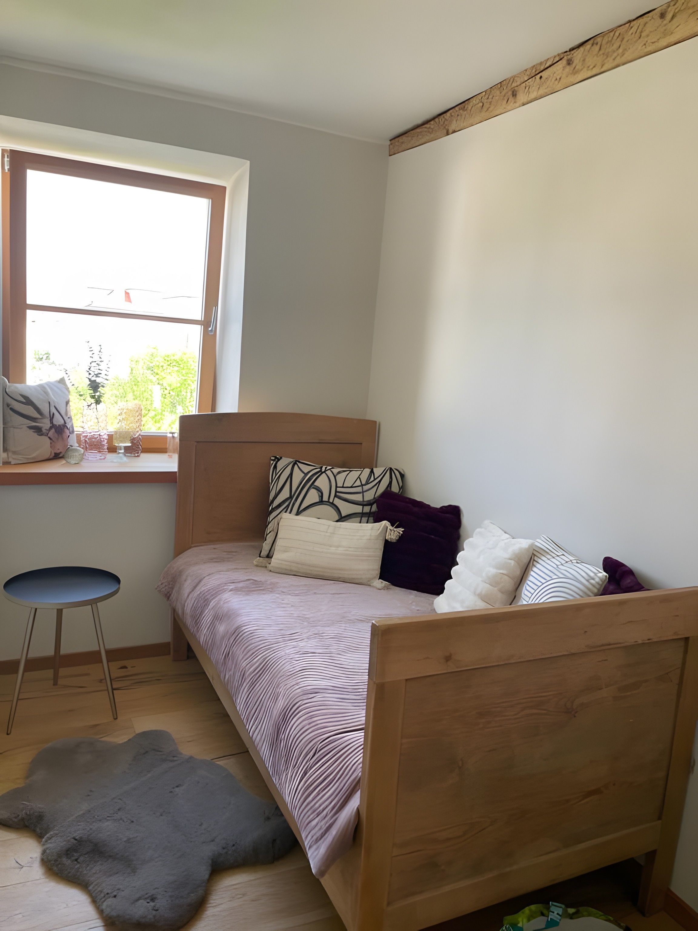 A small, single-bed room with a wooden headboard, a window with a view of greenery, a small stool, and a fur rug on the floor.
