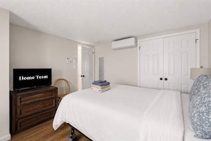 Un dormitorio con una televisión sobre una cómoda de madera, una cama con ropa de cama blanca, una pila de ropa doblada y una puerta.