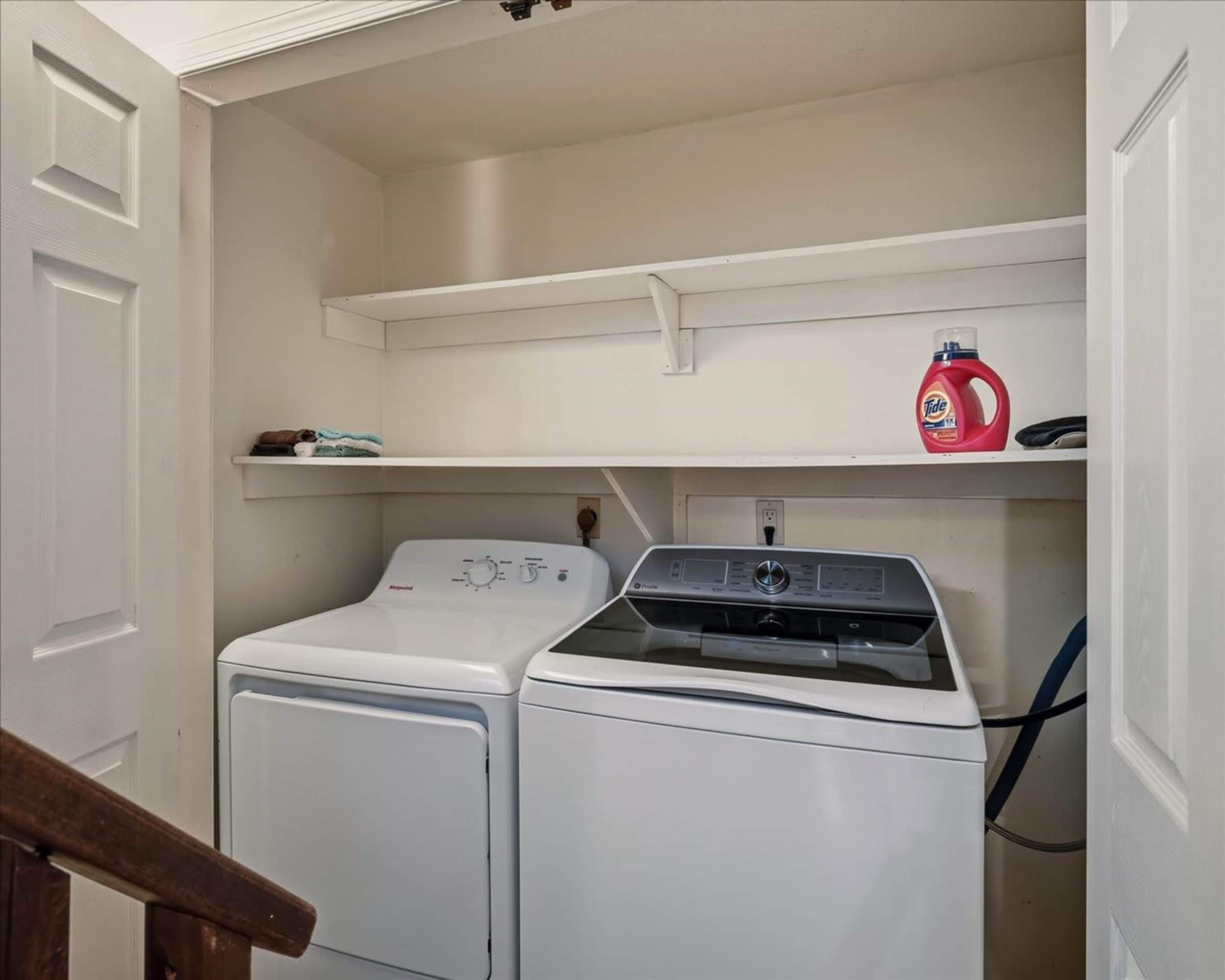 A laundry room with two front-loading washing machines, a shelf with folded towels, and a bottle of Tide detergent.