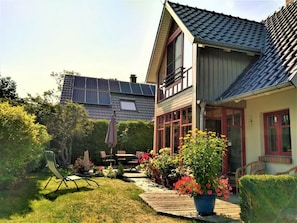 Una casa de dos pisos con techo inclinado, paneles solares, un patio con mobiliario exterior y un jardín con plantas en macetas.