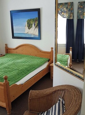 A bedroom with a bed, a wicker chair, a painting of cliffs, and a mirror reflecting the room. - Quisisana House, Apt. 15 (Göhren)