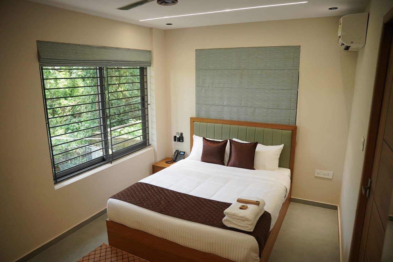 Hotelkamer met een bed, een raam met zonwering, een nachtkastje met telefoon en een wandairconditioning.