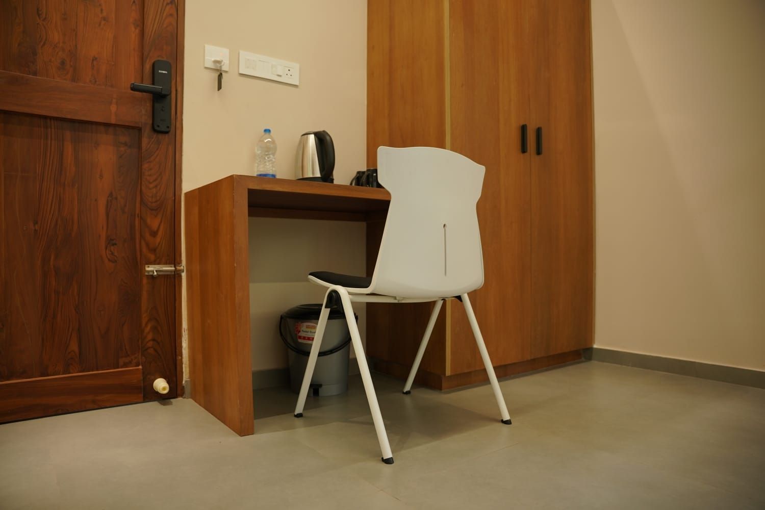 A room with a wooden wardrobe, a white chair, a desk with a kettle, and a trash bin.