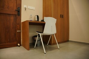 A room with a wooden wardrobe, a white chair, a desk with a kettle, and a trash bin.