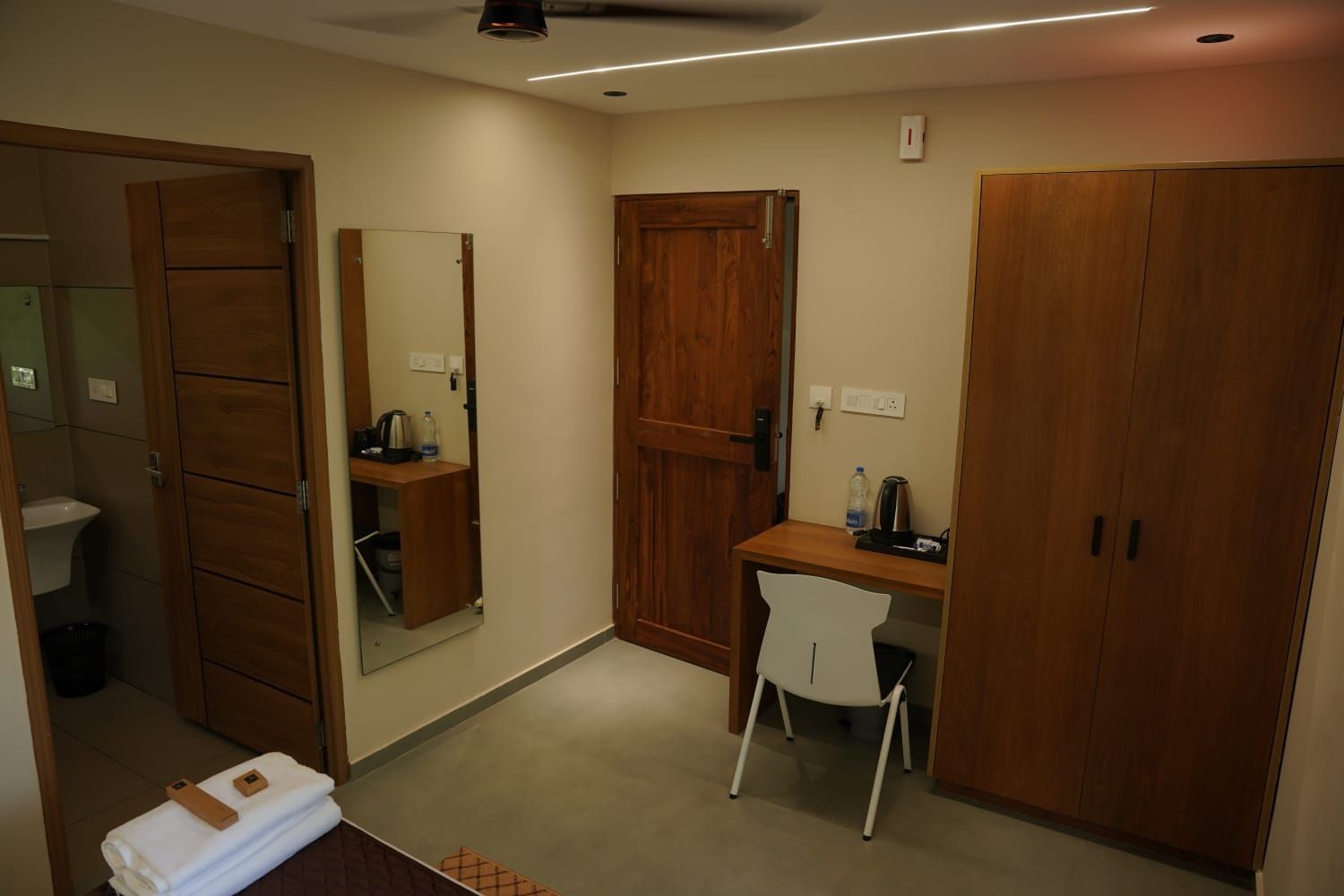 A hotel room with a wooden wardrobe, a desk with a chair, a mirror, and a small shelf with toiletries.