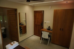 A hotel room with a wooden wardrobe, a desk with a chair, a mirror, and a small shelf with toiletries.