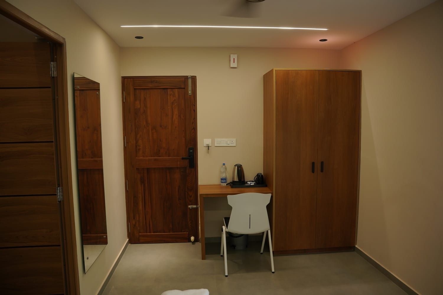 A room with a wooden wardrobe, a desk with a chair, and a small table with a water bottle and a kettle.