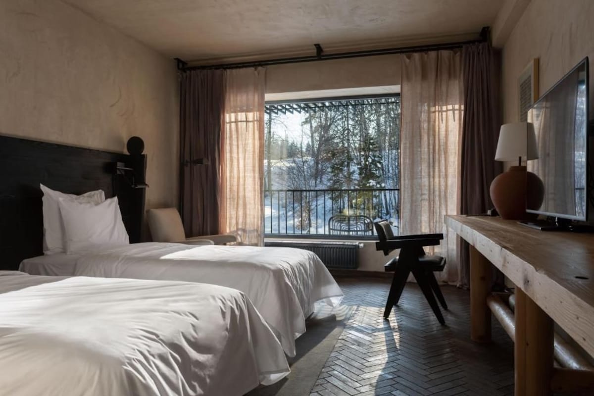 Une chambre d’hôtel avec deux lits, un bureau, une chaise, une fenêtre donnant sur des arbres et un balcon.