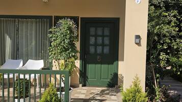Una entrada de edificio con una puerta verde, un pequeño jardín con plantas en macetas y una silla blanca en el exterior.
