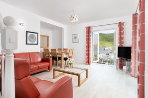 Ein modernes Wohnzimmer mit einem roten Sofa, einem Glastisch und einem Essbereich mit Holzkästen. Ein Fernseher ist an der Wand befestigt, und durch die Vorhänge ist eine grüne Landschaft zu sehen.
