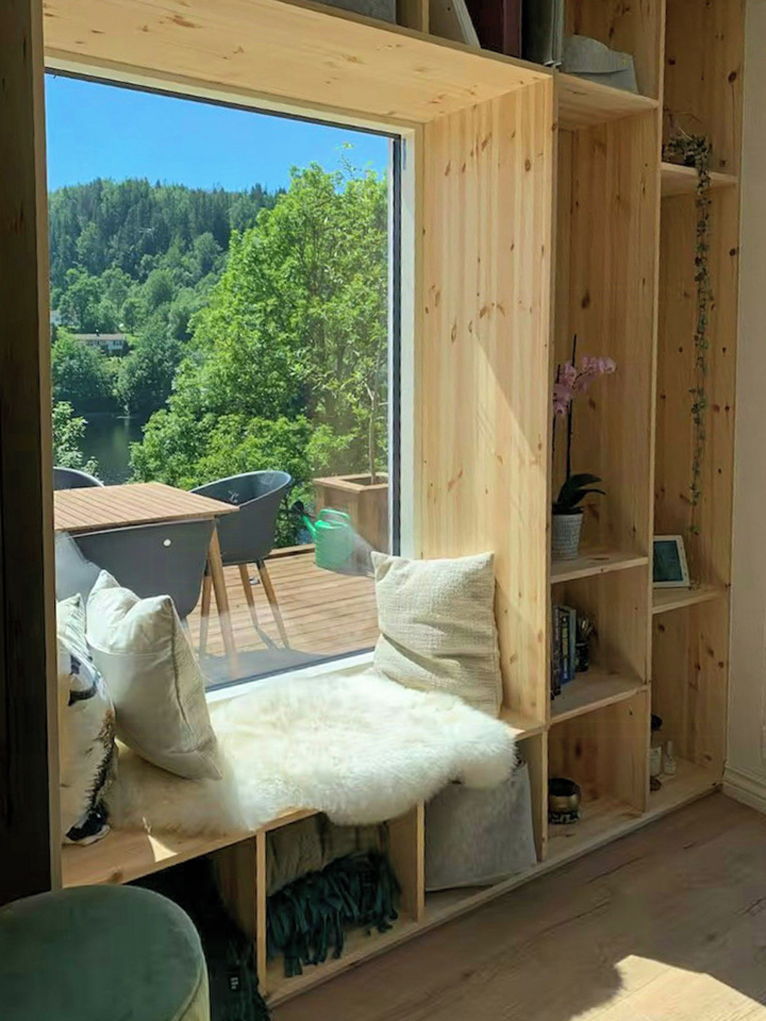 Ein Zimmer mit einem großen Fenster, Holzregalen, einer Sitzecke mit Kissen und Blick auf ein bewaldetes Gebiet.