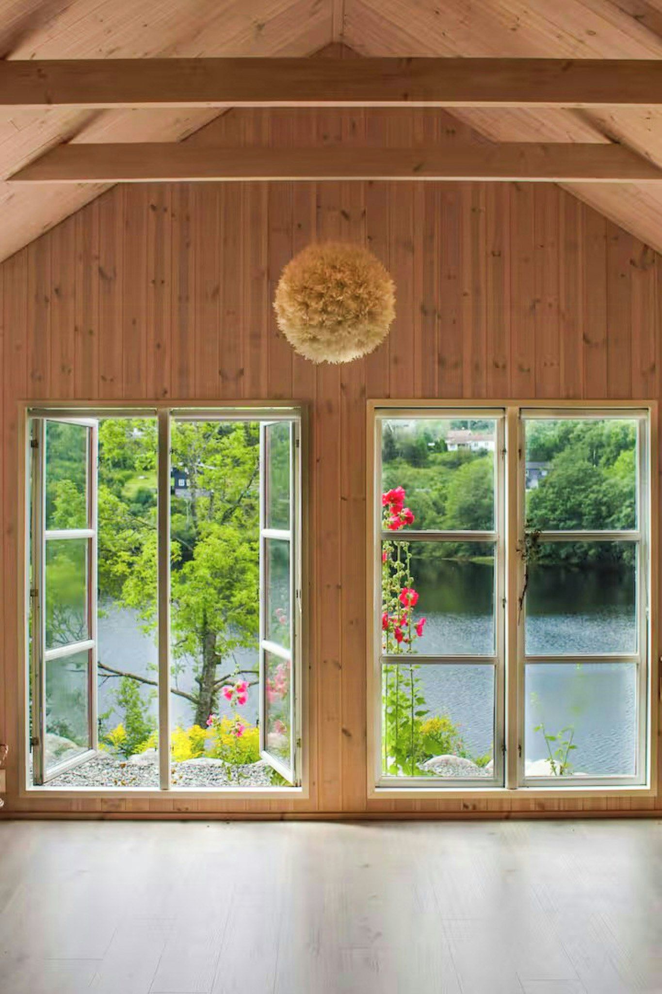 Una habitación con paredes de madera y un techo alto, con dos ventanas amplias que dan a un lago y vegetación.