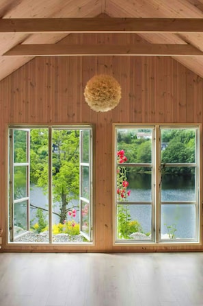 Una habitación con paredes de madera y un techo alto, con dos ventanas amplias que dan a un lago y vegetación.