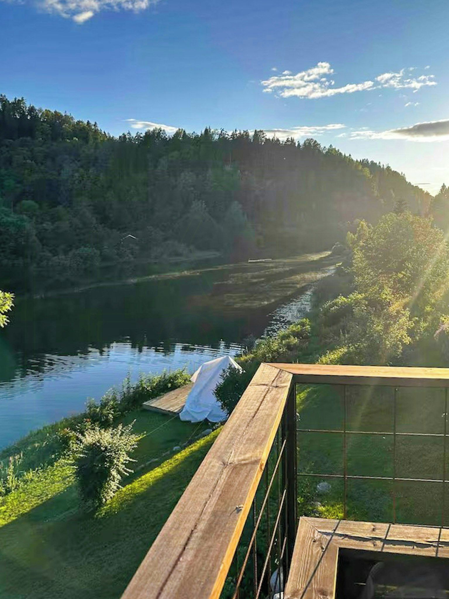 A riverside view with a wooden deck overlooking a river, surrounded by greenery and hills.
