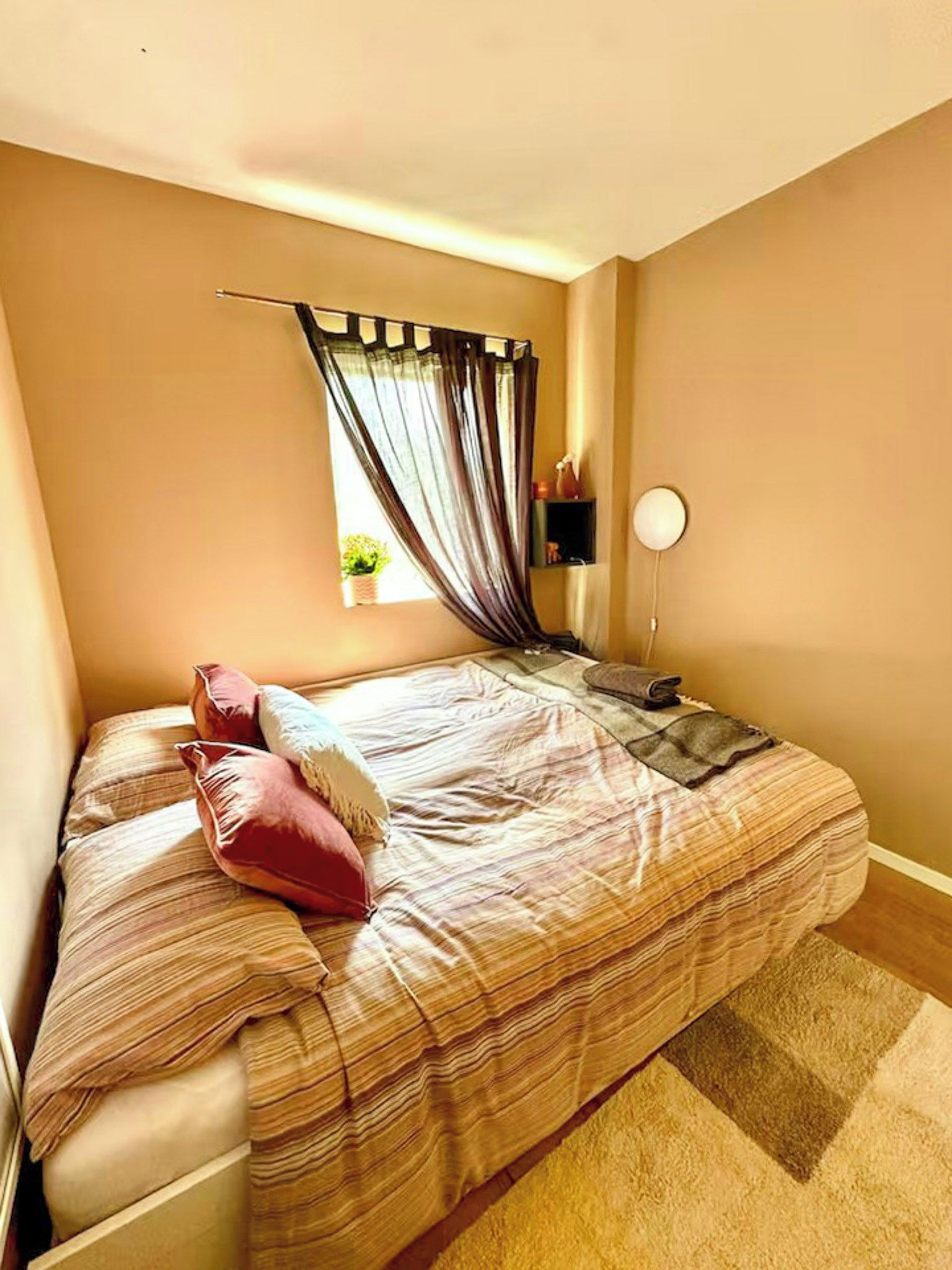 A bedroom with a bed, pillows, a bedside table, a lamp, and a window with curtains.