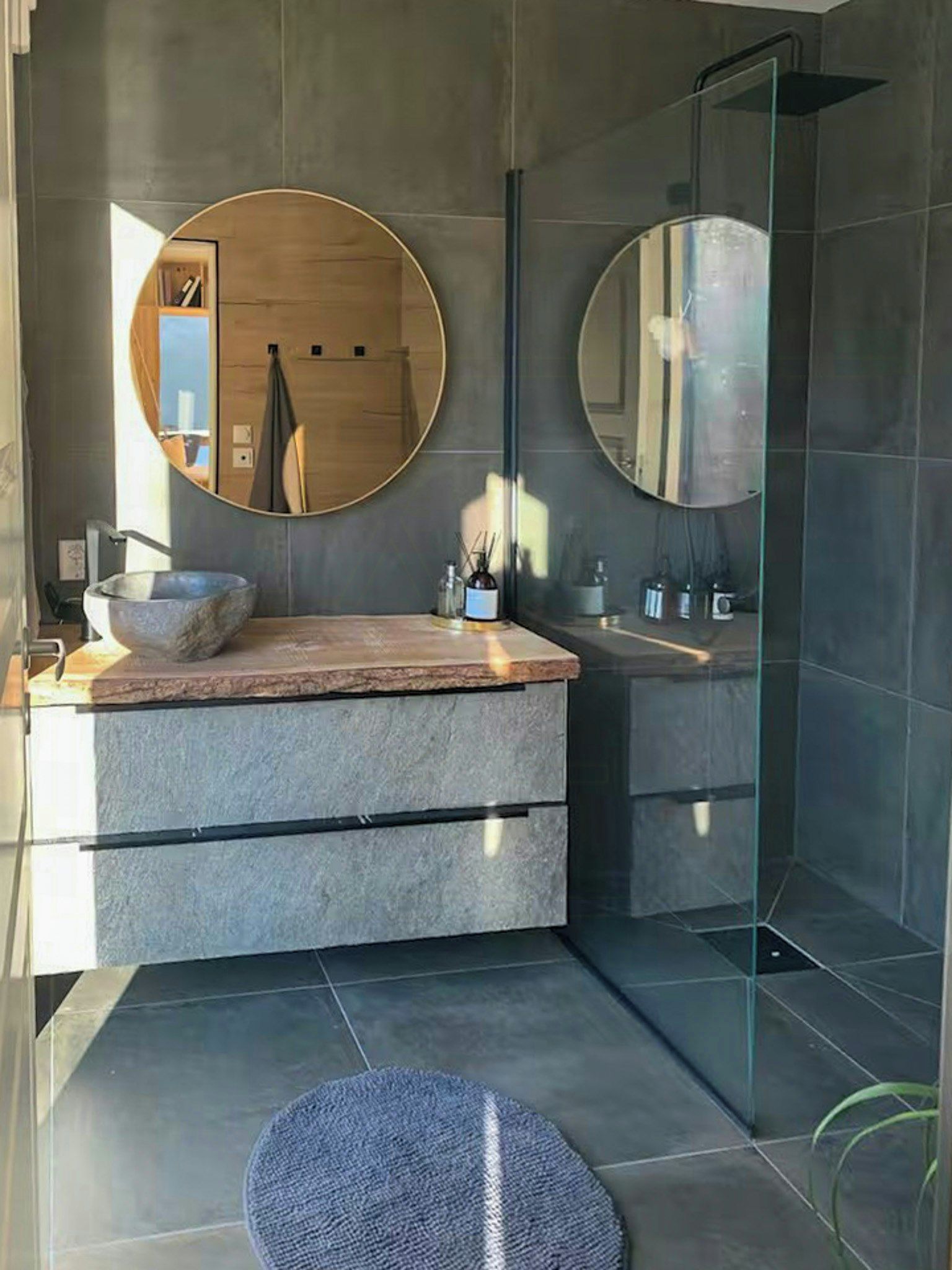 Ein modernes Badezimmer mit einem Naturstein-Waschtisch, zwei runden Spiegeln, einer Duschkabine aus Glas und einem runden Teppich auf dem Boden.