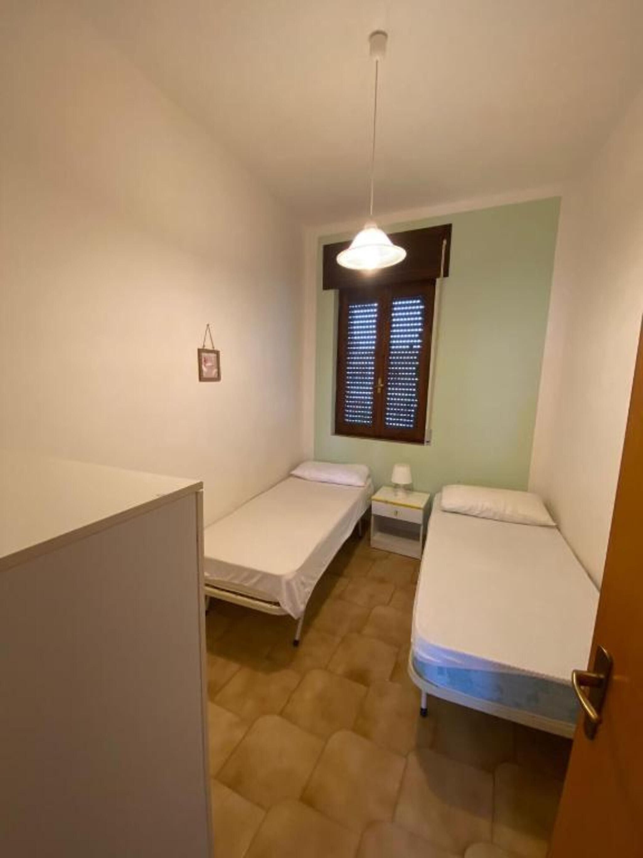 A small, compact room with two single beds, a bedside table with a lamp, a hanging light fixture, and a window with blinds.
