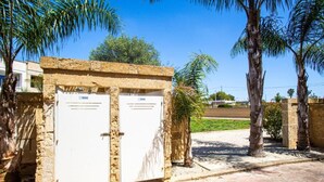 Um edifício com duas portas brancas, uma parede de pedra e um espaço pavimentado com palmeiras.