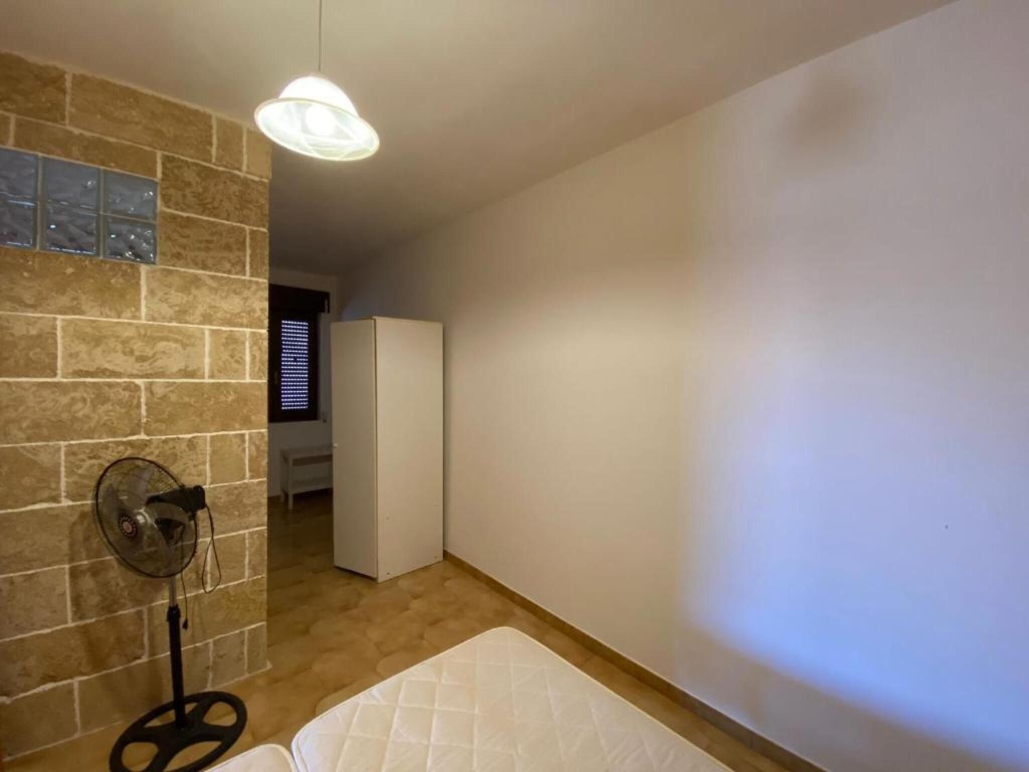 A room with a brick wall, a white ceiling fan, a white mattress, and a door leading to another room.