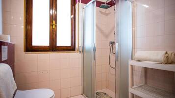 A bathroom with a glass-enclosed shower, a toilet, and a shelf with towels.
