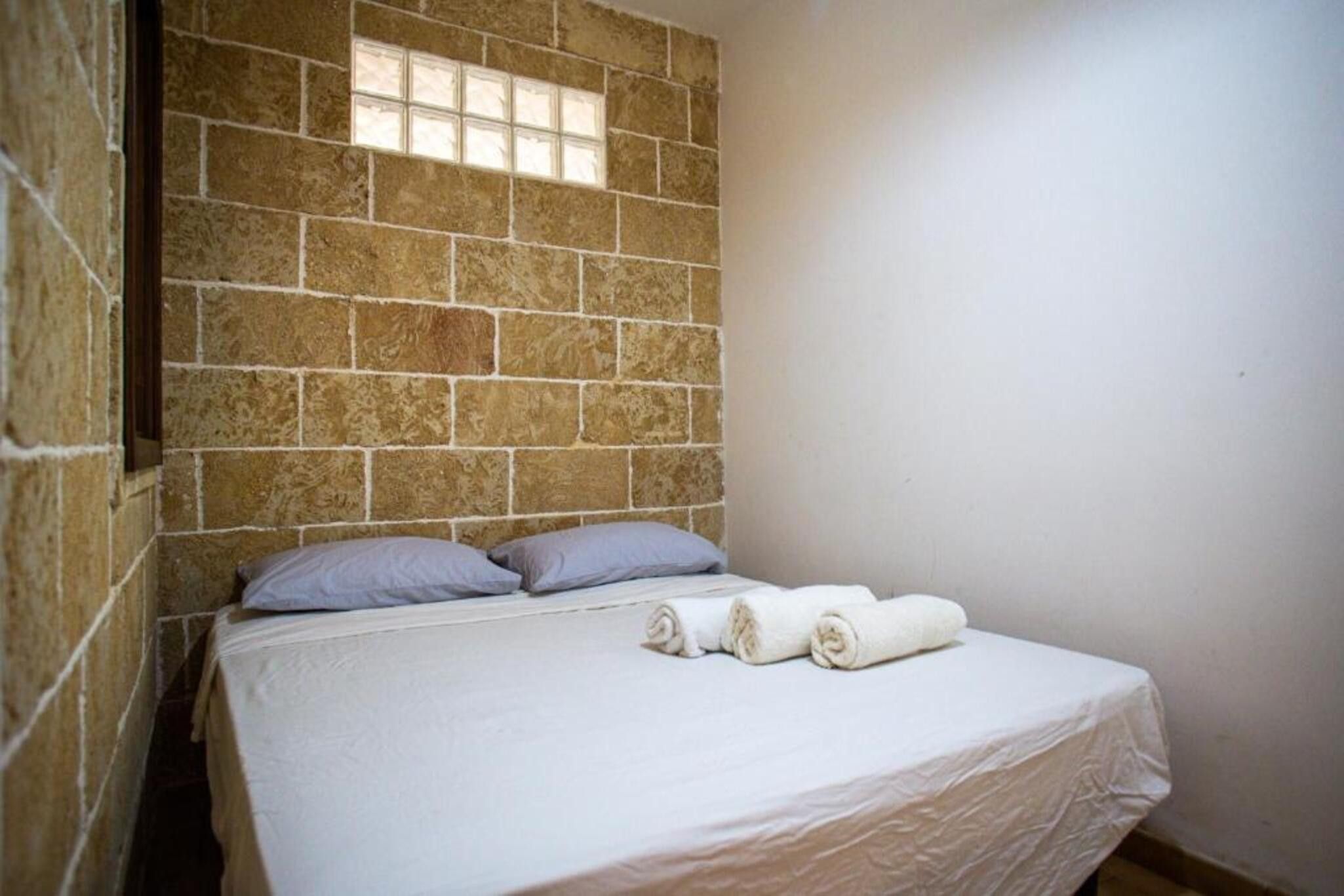 Ein Einzelbett mit weißer Matratze, zwei Kissen und zwei zusammengerollten Handtüchern in einem Zimmer mit Steinwänden und einem Fenster.