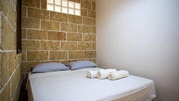 Ein Einzelbett mit weißer Matratze, zwei Kissen und zwei zusammengerollten Handtüchern in einem Zimmer mit Steinwänden und einem Fenster.