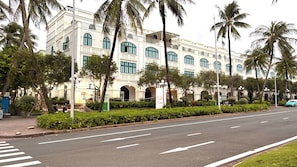 A street view with a large building featuring arched windows and a balcony, surrounded by palm trees and a crosswalk. - Diamond Suite Lotus Village (Phu Quoc)