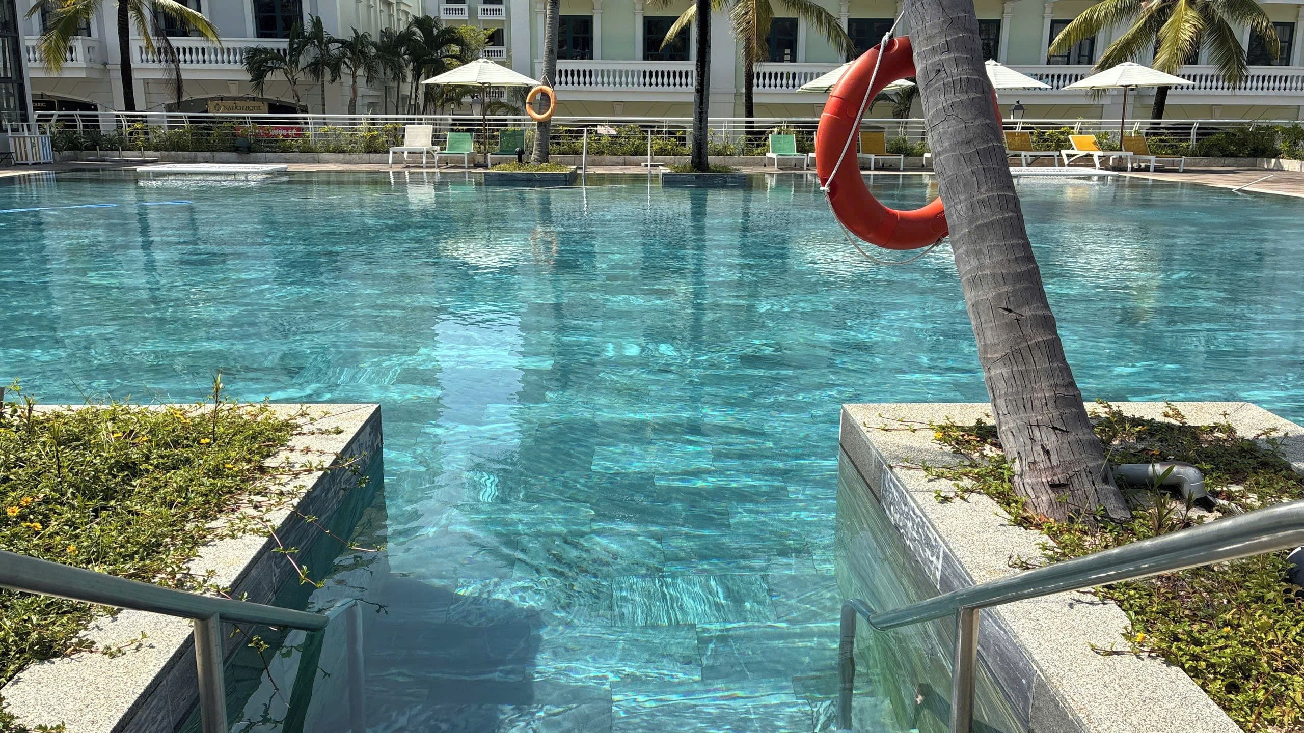 Piscina cristalina com um salva-vidas preso a uma palmeira, rodeada por espreguiçadeiras e guarda-sóis.