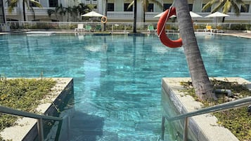 Piscina cristalina com um salva-vidas preso a uma palmeira, rodeada por espreguiçadeiras e guarda-sóis.