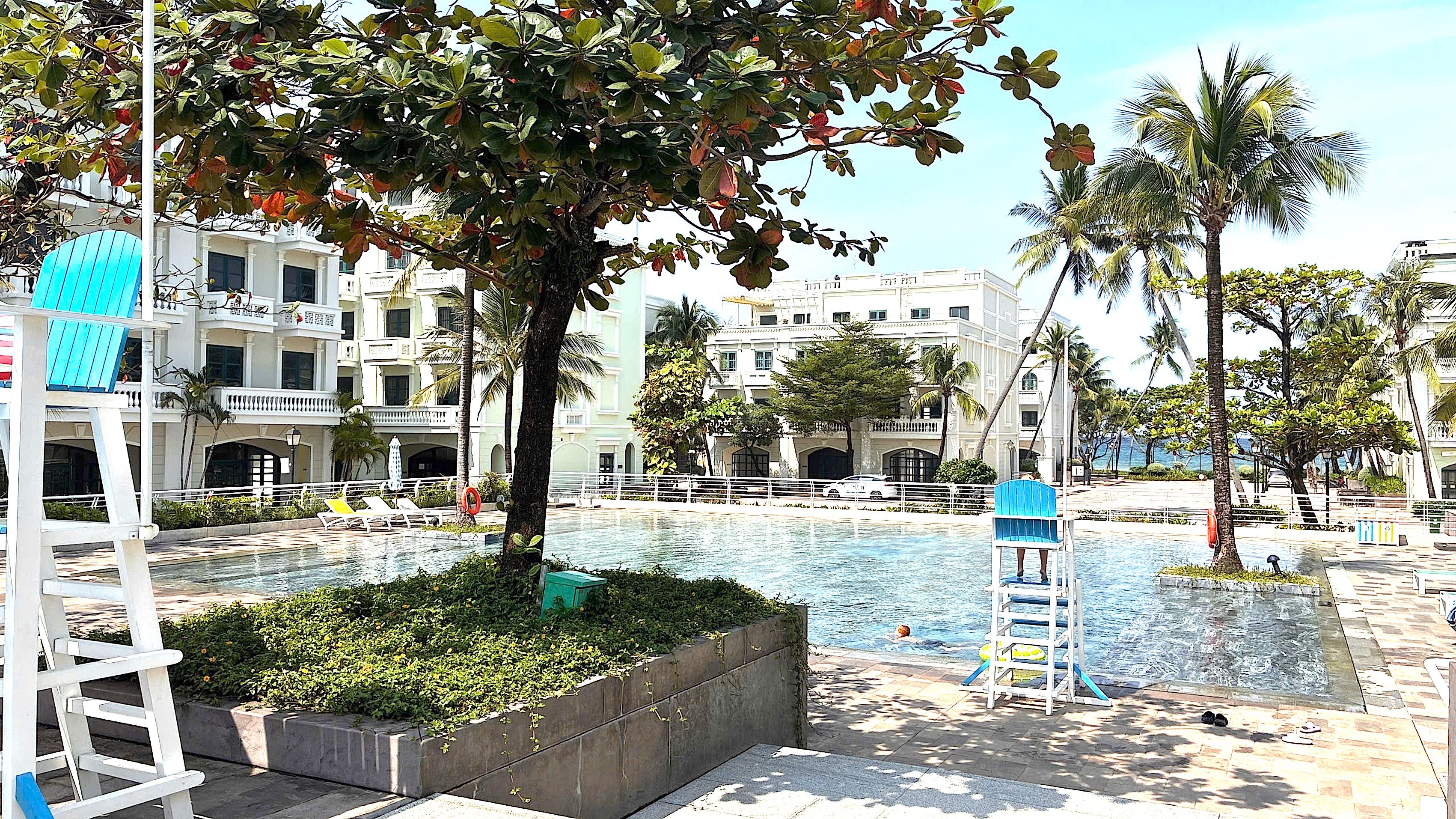 Zona de piscina con sillones de descanso, puesto de socorrista y un edificio con varios balcones al fondo.