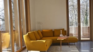 A modern living room with a mustard-colored sofa, a small round table with a bottle and glasses, and a large window with a view of trees. - bel Etage Avenue in the Parkchalet (Potsdam)