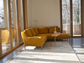 En moderne stue med en sennepsgul sofa, et lille rundt bord med en flaske og glas, samt et stort vindue med udsigt til træer.