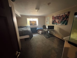 Quarto com cama, escrivaninha com laptop, janela e pôsteres nas paredes.