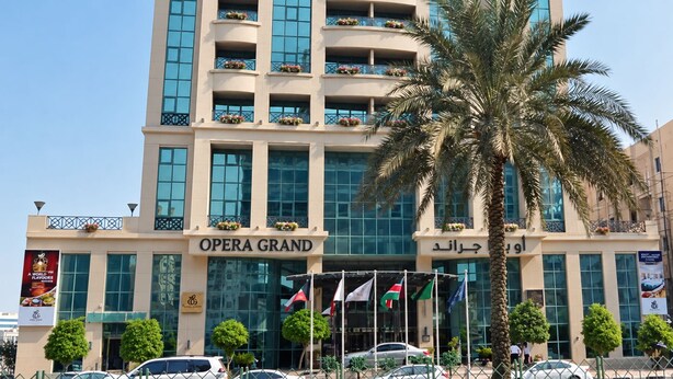 A multi-story building with balconies, a palm tree in the foreground, and flags at the entrance. - Opera Grand Hotel Deira (Dubai)