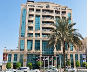 A multi-story building with balconies, a palm tree in the foreground, and flags at the entrance. - Opera Grand Hotel Deira (Dubai)
