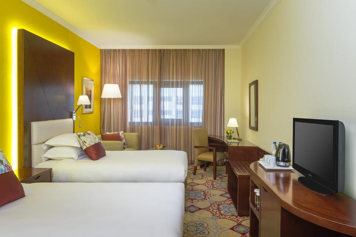 Habitación de hotel con dos camas, un televisor, un escritorio con lámpara y una ventana con cortinas.