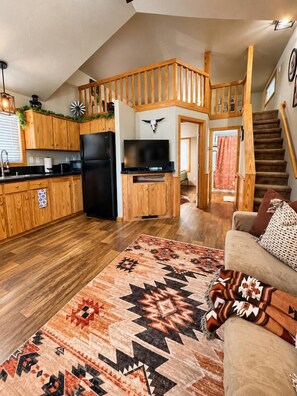 A spacious living area with a wooden staircase, a kitchen with a black refrigerator, and a patterned rug. - Lake Travis Cabin Retreat + Pool, Hot Tub, Gym (Austin)