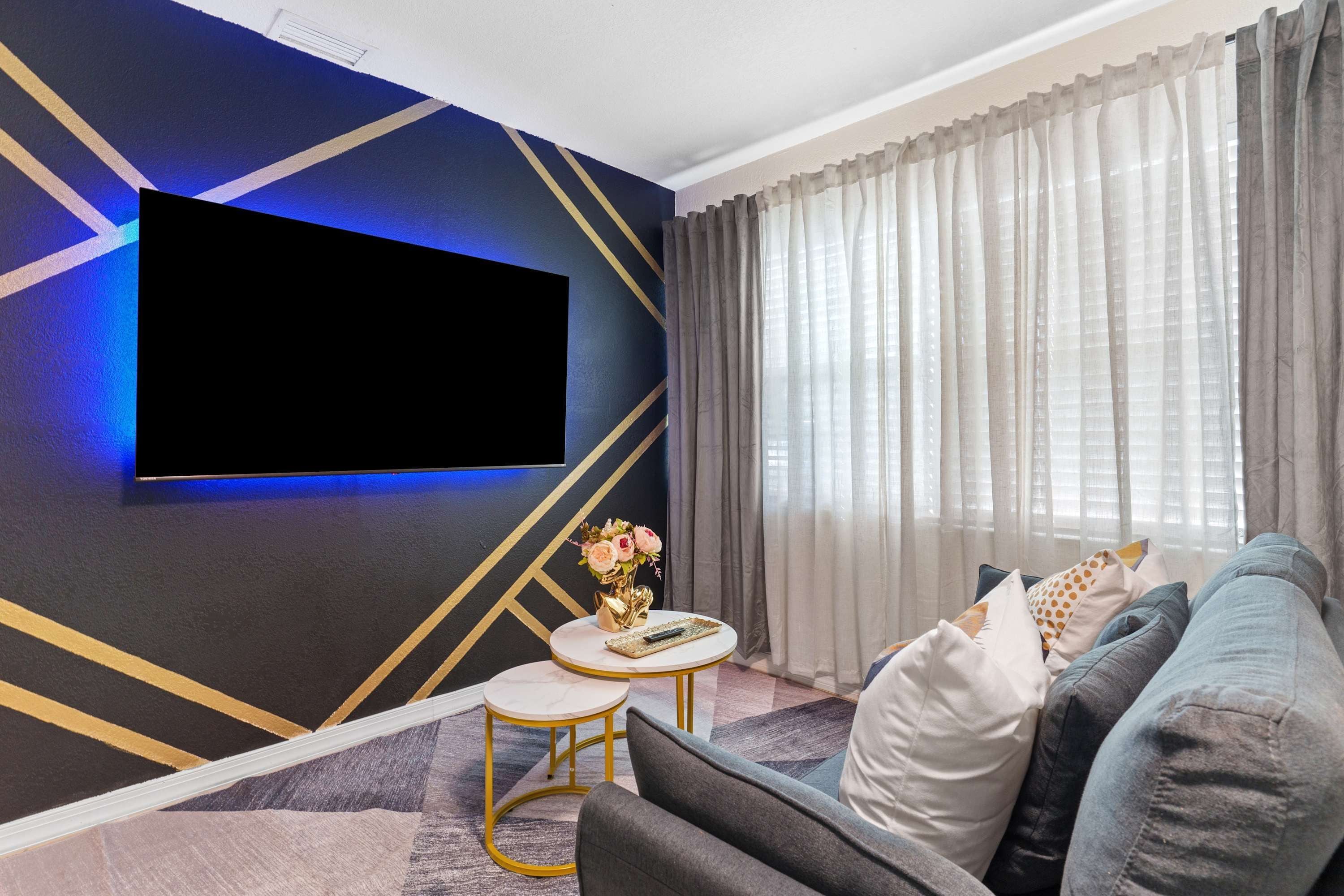 A modern living room with a flat-screen TV mounted on the wall, a round coffee table with a vase of flowers, and a sofa with decorative pillows.