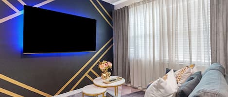 A modern living room with a flat-screen TV mounted on the wall, a round coffee table with a vase of flowers, and a sofa with decorative pillows.