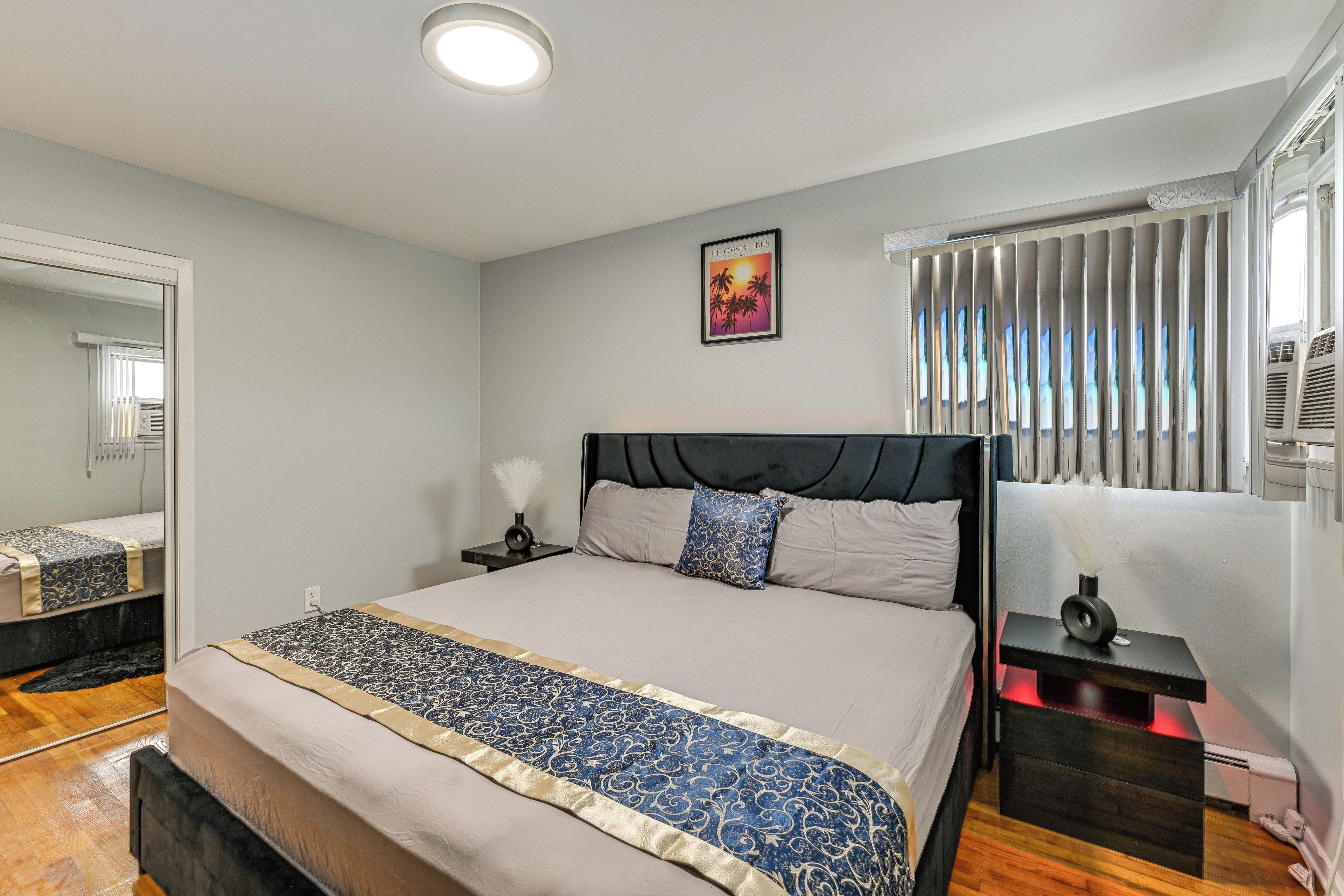 Un dormitorio con cama, mesita de noche, espejo, ventana con persianas y aire acondicionado de pared.