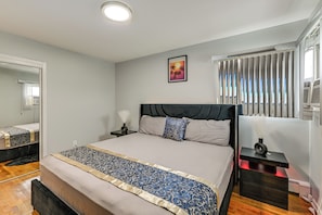 A bedroom with a bed, a nightstand, a mirror, a window with blinds, and a wall-mounted air conditioner. - 3 Mi to Jfk: Convenient NYC Base w/ Shared Patio (Jamaica)