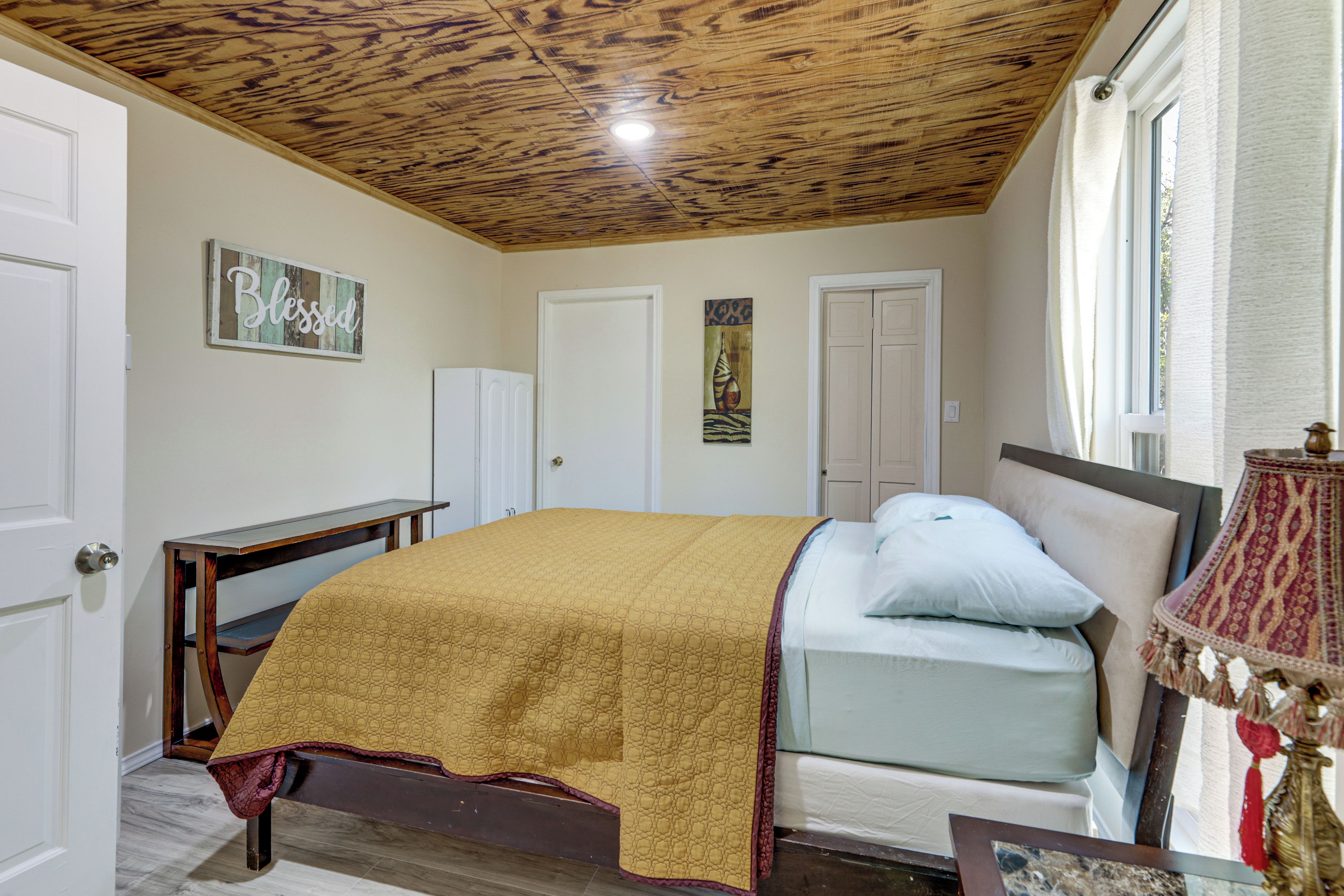 A bedroom with a wooden ceiling, a bed with a yellow cover, a bedside table, a lamp, and a painting on the wall.