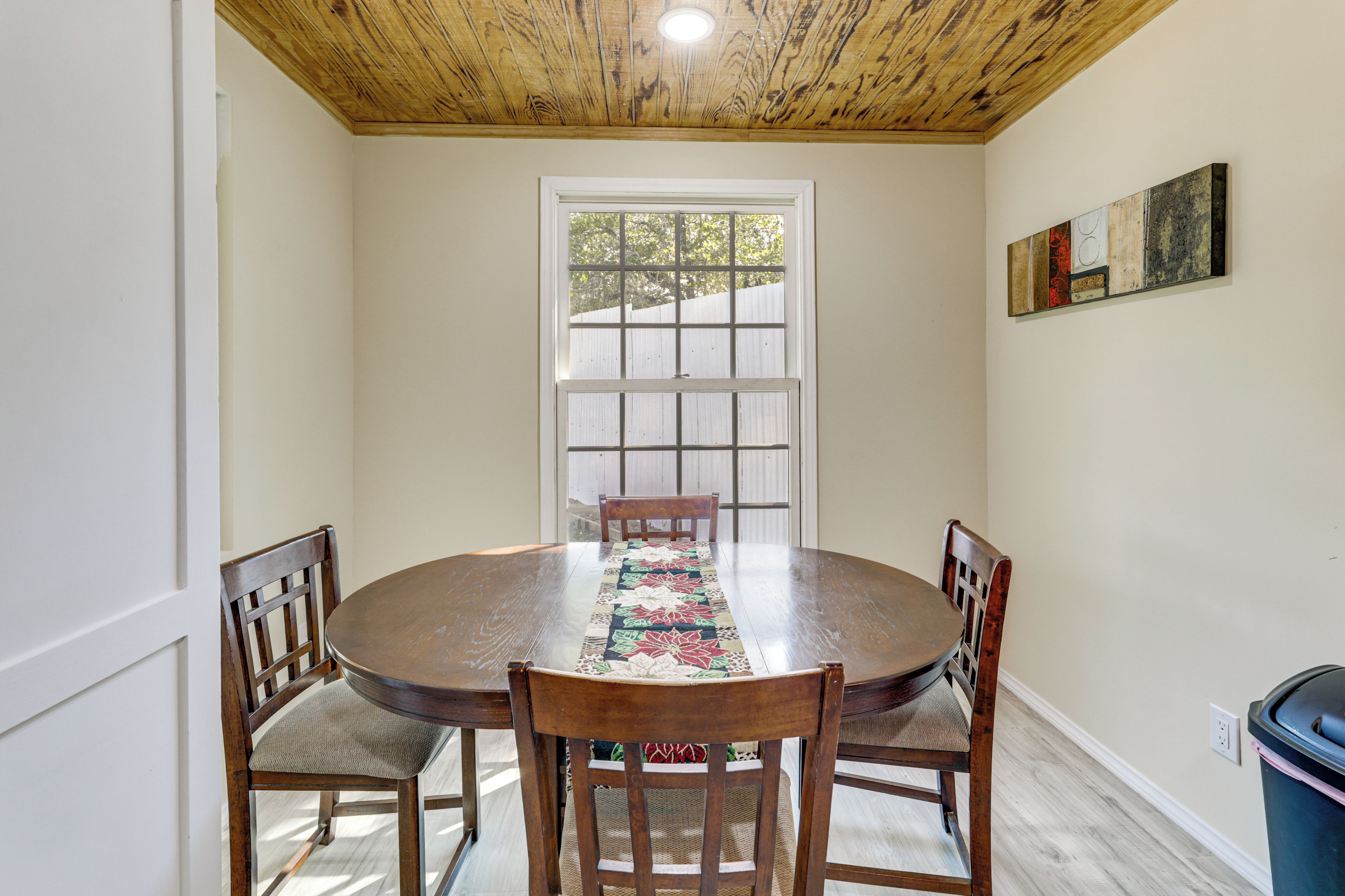 A dining room with a wooden table, four chairs, a window with blinds, and a painting on the wall.