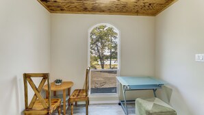 Quarto com teto de madeira, uma mesa com cadeiras, um banquinho e uma janela com vista para árvores.