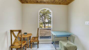 A room with a wooden ceiling, a table with chairs, a small stool, and a window with a view of trees.