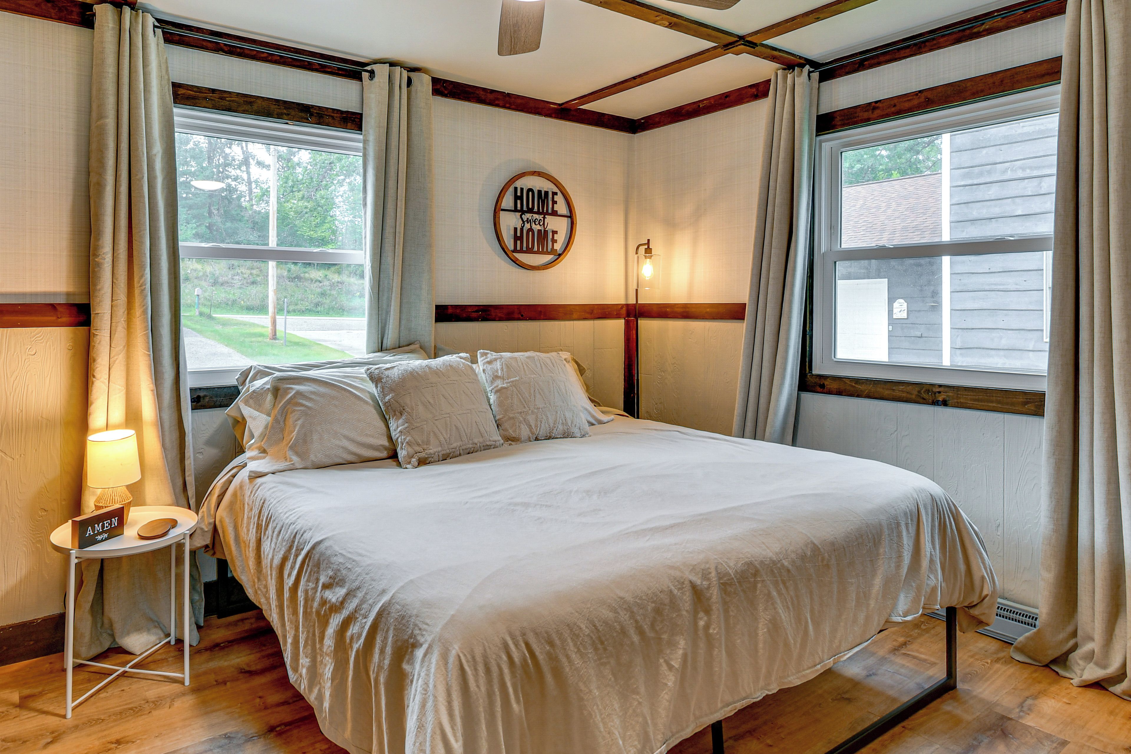 A bedroom with a bed, a bedside table with a lamp, a wall clock, and a window with curtains.
