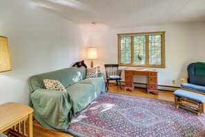 A cozy room with a green sofa, a wooden desk, a chair, a patterned rug, and a window with a view of trees. - Gateway to Acadia Nat'l Park! Peaceful Home (Ellsworth)