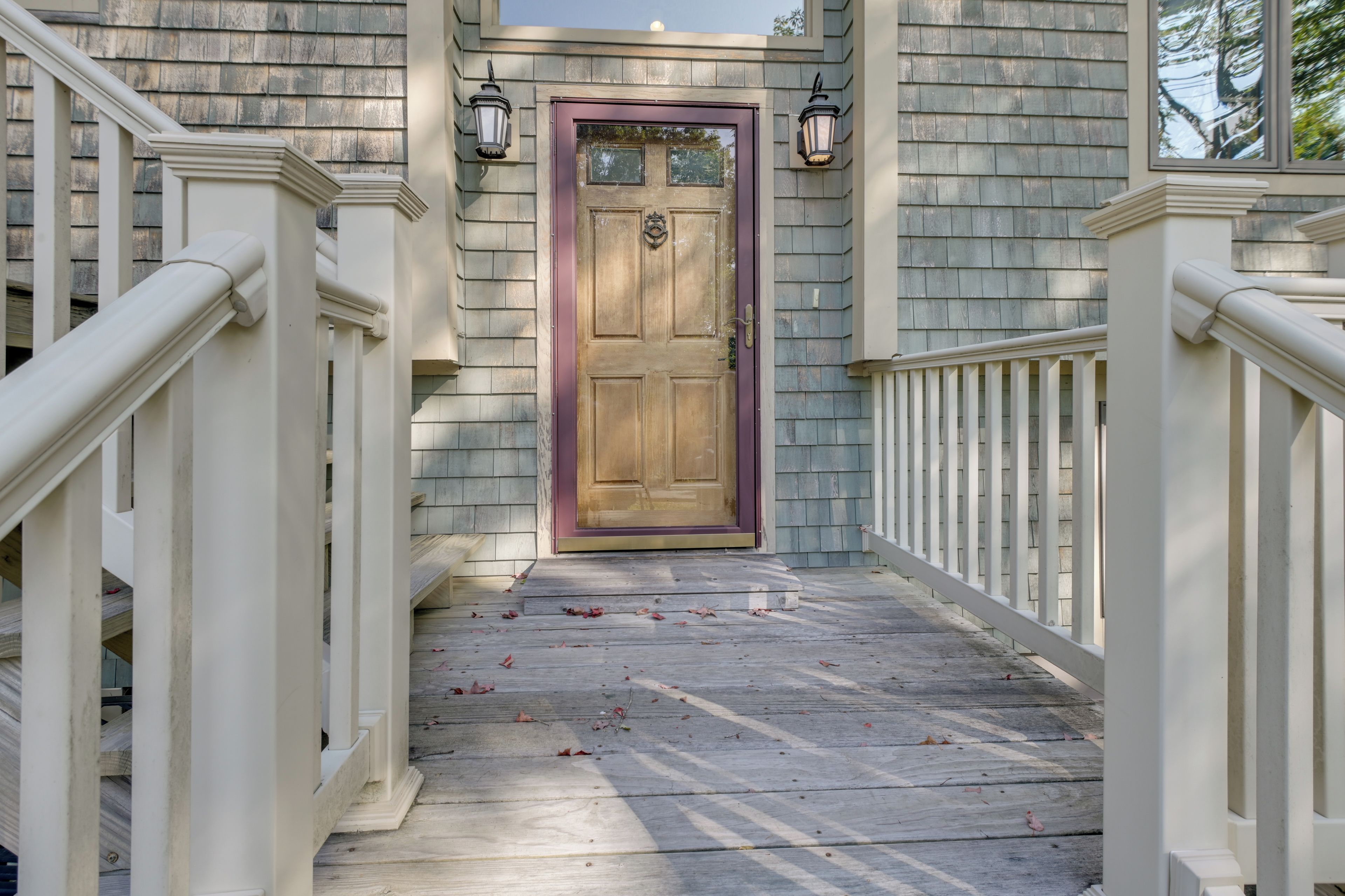 A wooden front door with a glass panel, flanked by white railings and two wall-mounted lanterns.