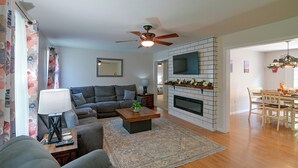 A living room with a sectional sofa, a coffee table, a fireplace, a TV mounted on a brick wall, and a dining area with a table and chairs. - Private Yard: Fire Pit & Cornhole! Moorefield Gem (Moorefield)